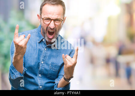 Mittleres Alter hoary älterer Mann mit Brille über isolierte Hintergrund schreien mit verrückten Ausdruck tun Rock Symbol mit hands up. Musik Star. Schwere Stockfoto