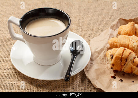 Tasse schwarzen Kaffee mit Löffel in Sacktuch, frische Croissants mit Schokolade auf Geschenkpapier Stockfoto