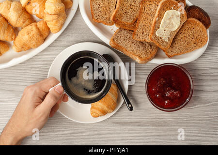 Blick von oben auf die männliche Hand, die eine Tasse schwarzen Kaffee am Tisch mit Croissant, Brot und Erdbeermarmelade in der Schüssel. Nahrungsmittel und Getränke zum Frühstück Stockfoto