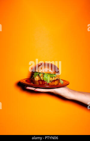 Hand, Chicken Burger auf rote Platte gegen orange hinterlegt. Stockfoto