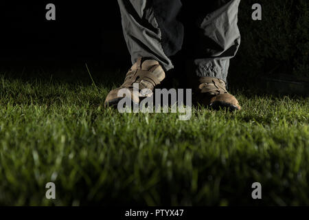 Niedrigen Winkel, Night Shot des Mannes Fuß in der Ausbilder, beleuchtet, stehend auf Gras im Garten. Der froschperspektive der männlichen Schuhe & Unterschenkel Hose in Dunkel. Stockfoto