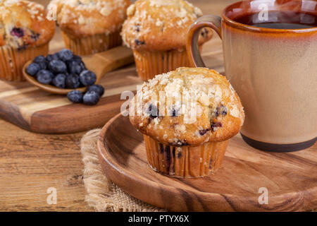 Blueberry Muffin und Tasse Kaffee auf einer Holzplatte mit Blaubeeren und Muffins im Hintergrund Stockfoto