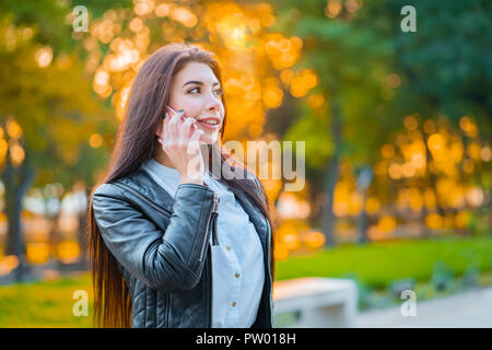 Business woman mit Smartphone in der Nähe im Herbst Park im Abendlicht. Mädchen haben Gespräch mit Handy. Schönen kaukasischen junge Frau talkin Stockfoto