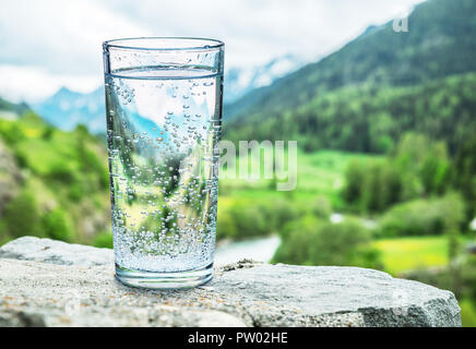 Glas Wasser auf den Stein. Verschwommen Schnee Berge Tops und grüne Wälder im Hintergrund, als Symbol für Frische und Reinheit. Stockfoto