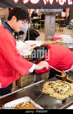 Japanischer Mann im traditionellen roten happi Jacke, bei Yakisoba takeaway Kochen gebratene Buchweizennudeln auf Warmhalteplatte während des Festivals. Stockfoto