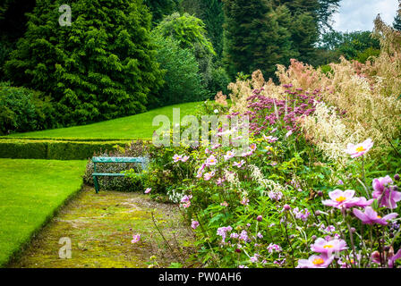 Alte Holz garten Sitz, Sommerzeit, von bunten Blumen in voller Blüte neben einem gepflegten grünen Rasen umgeben, Große grüne Bäume im Hintergrund. Stockfoto