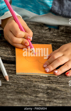 Nahaufnahme auf childs Hand zeichnen ein Herz auf Papier mit farbigen rosa Bleistift über Verwitterten Holztisch. Stockfoto