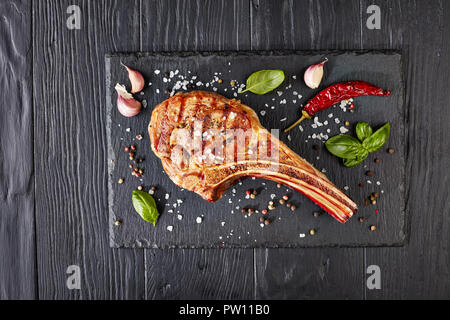 Close-up von gegrilltem Tomahawk Steak oder Cowboy Rindersteak auf schwarzem Schiefer mit Knoblauch, frischem Basilikum, Ansicht von oben, flach Stockfoto
