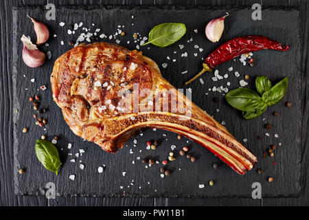 Close-up von gegrilltem Tomahawk Steak oder Cowboy Rindersteak auf schwarzem Schiefer mit Knoblauch, frischem Basilikum, Ansicht von oben, flach Stockfoto