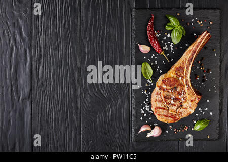 Gebratene tomahawk Steak oder Cowboy Rindersteak auf schwarzem Schiefer mit Knoblauch, frischem Basilikum, Ansicht von oben, flach Stockfoto