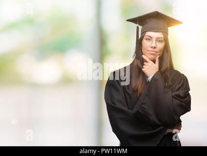 Junge Hispanic Frau tragen graduierte Cap und einheitliche schaut zuversichtlich in die Kamera mit einem Lächeln mit verschränkten Armen und Hand am Kinn angehoben. Denken Stockfoto