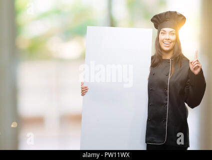 Junge hispanic Koch Frau Koch einheitliche Holding banner überrascht mit einer Idee oder Frage Zeigefinger mit glücklichen Gesicht, Nummer eins Stockfoto
