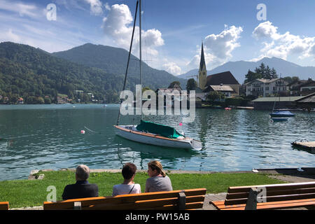 Blick auf den Tegernsee und Rottach Egern mit den Wallberg. Im Vordergrund Menschen auf einer Bank sitzen. Ein Segelboot liegt im Wasser, | Verwendung weltweit Stockfoto