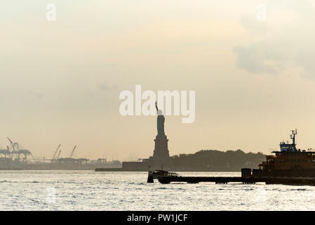 10-2018 Manhattan, New York. Die Freiheitsstatue in der Dämmerung, mit der Staten Island Ferry an der rechten, und Kräne und Docks von New Jersey auf der linken Seite. Foto: Stockfoto