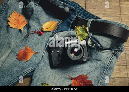 Zusammensetzung der Blätter im Herbst, retro Kamera und Blue Denim Jeans auf Holz- Hintergrund. Ansicht von oben, flach. In warmen Farbtönen. Konzept der Herbst Foto schießt. Stockfoto