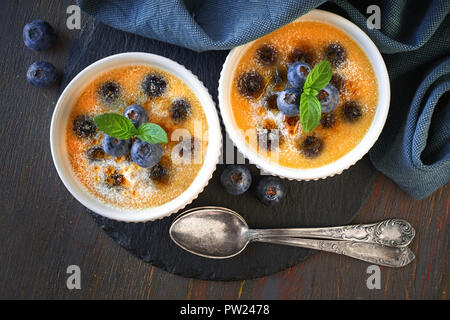 Crème Brûlée (Creme brulee, Gebrannte Creme) mit Blueberry in Pulverform mit Zucker auf dunklen Holzmöbeln im Landhausstil Hintergrund Stockfoto