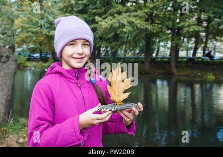 Mädchen in blau und magenta Jacke holding Holz handgefertigt kleines Spielzeug floss mit eichenlaub als Segel im Freien in der Nähe des Flusses auf düsteren Herbst Tag Stockfoto