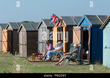 Die Küste der Insel Hayling mit Kiesstrände und bunten Badekabinen, Hampshire, UK. Drei Personen, die vor einer Strandhütte sitzen. Stockfoto