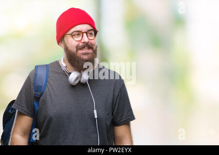 Junge hipster Mann mit roten wolle Gap und Rucksack über isolierte Hintergrund entfernt, mit einem Lächeln auf dem Gesicht zu Seite, natürlicher Ausdruck. Lachend con Stockfoto