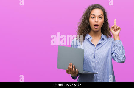 Junge Hispanic woman holding Computer Laptop mit einer Idee oder Frage Zeigefinger mit glücklichen Gesicht überrascht, Nummer eins Stockfoto