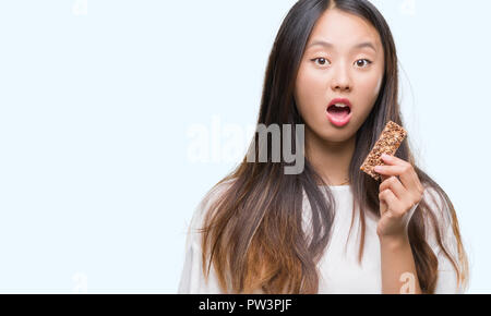 Junge asiatische Frau Schokolade essen energetische Bar über isolierte Hintergrund im Schock Angst mit einer Überraschung konfrontiert, ängstlich und aufgeregt mit Furcht expressio Stockfoto