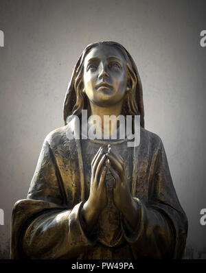 Heilige Maria Statue aus Bronze, die betet mit gefalteten Händen, im Hintergrund eine graue Wand. Er kann für Veranstaltungen, Konzepte und Hintergründe verwendet werden. Stockfoto