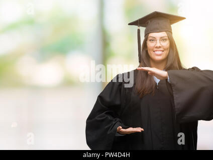 Junge Hispanic Frau tragen graduierte Cap und einheitliche gestikulierte mit Händen zeigen große und große Zeichen, Symbol. Lächelnd mit Blick auf den Ca Stockfoto
