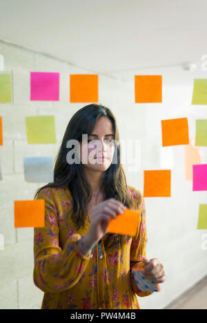 Geschäftsfrau klebt Haftnotizen an Fenster im kreativen Büro durch Glas Stockfoto