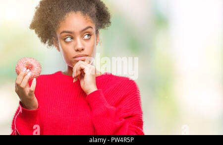 Junge afro-amerikanische Frau essen Donut über isolierte Hintergrund ernstes Gesicht denken über Frage, sehr verworrene Vorstellung Stockfoto