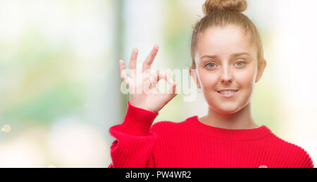 Junge blonde Frau mit Brötchen und roten Pullover Lächeln positive tun ok Zeichen mit der Hand und der Finger. Erfolgreiche Ausdruck. Stockfoto