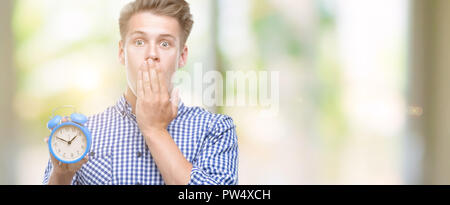 Jungen gutaussehenden blonden Mann holding Wecker decken den Mund mit der Hand mit Schande für den Fehler, den Ausdruck von Angst erschüttert, die in der Stille Angst, geheime conce Stockfoto