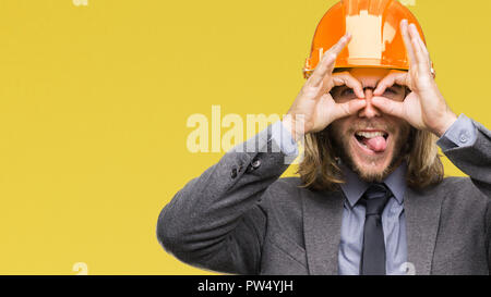 Junge schöne architec Mann mit langem Haar zu tragen Schutzhelm über isolierte Hintergrund tun ok Geste wie Fernglas Zunge heraus, Augen l Stockfoto