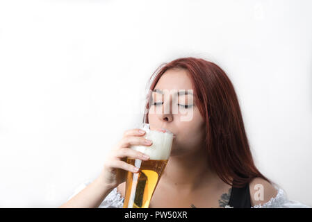 Eine junge, hübsche Frau mit langen roten Haaren in einem traditionellen deutschen Korsett Getränke eine goldene Bier aus einem Glas Stockfoto