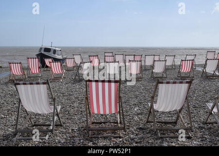 Reihen von leeren roten und weißen Liegestühlen am Strand mit Blick aufs Meer und den blauen Himmel im Hintergrund, kleinen blauen und weißen Boot auf der linken Seite. Stockfoto