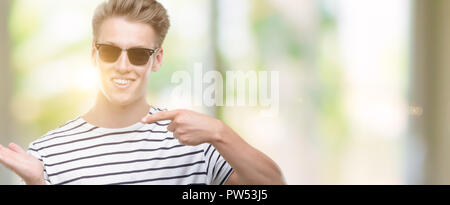 Jungen gutaussehenden blonden Mann, der sunglasess sehr glücklich zeigte mit Hand und Finger Stockfoto