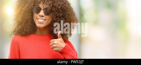 Afrikanische amerikanische Frau mit Sonnenbrille lächelte breit Daumen hoch, Geste, die Kamera, den Ausdruck von wie und Genehmigung Stockfoto