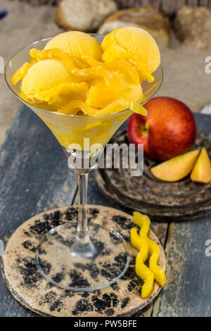 Glas Schale mit Mango pfirsich Obst Eis, Sorbet Kugeln auf Stein plank auf sandigen Strand serviert Stockfoto