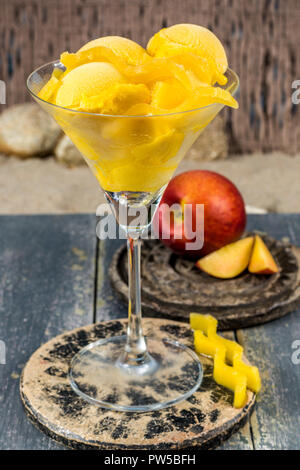 Glas Schale mit Mango pfirsich Obst Eis, Sorbet Kugeln auf Stein plank auf sandigen Strand serviert Stockfoto