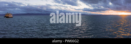 Panorama mit Sonnenuntergang über dem See Wasser und einzelne Boot segeln mit Passagieren, auf dem Bodensee in der Stadt Friedrichshafen, Deutschland Stockfoto