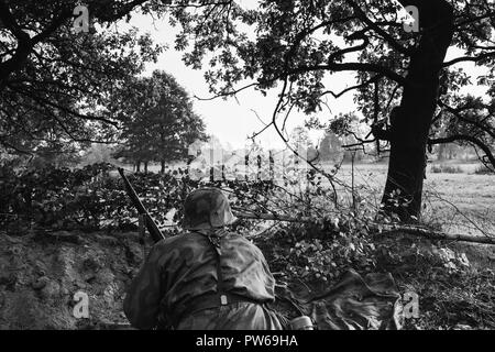 Re-Enactor gekleidet, wie Deutsche Wehrmacht Infanterie Soldat im Zweiten Weltkrieg versteckt Sitzen mit Gewehr Waffe in einen Hinterhalt in Graben im Herbst Wald. Phot Stockfoto