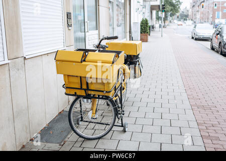 Traditionelle deutsche gelb Bike für Mail Lieferung ist auf der Straße in der Nähe des Hauses geparkt, während der briefträger die Post trägt. Post in Deutschland. Stockfoto