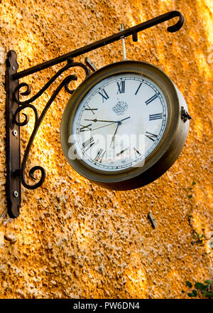 Alte antike Uhr mit römischen Ziffern, die an rostigen Klammern außen gegen die leuchtend orange farbige Wand hängen. Stockfoto