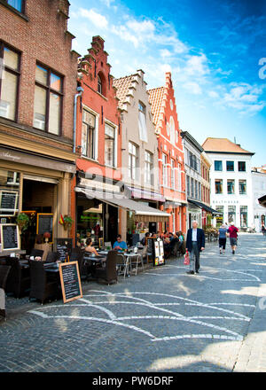 Ein elegant gekleideter Mann, der durch eine ziemlich gepflasterte Straße geht, bunte Giebelhäuser auf der einen Seite mit Cafés und kleinen Restaurants, historische Altstadt brügge Stockfoto