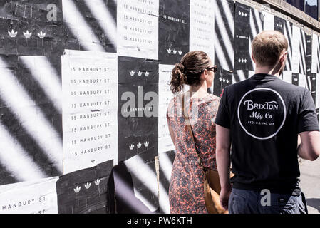 Paar von hinten zu Fuß auf einer Straße in Brixton neben einer Wand in schwarz-weiss Poster abgedeckt fotografiert. Brixton, Lambeth, South London, Großbritannien Stockfoto