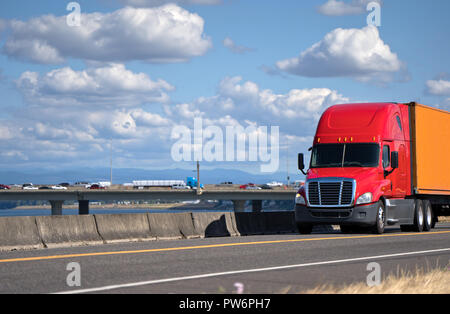 Kraftvolle rot Big Rig Semi Truck mit Container auf Flachbett Auflieger für den Transport von kommerziellen Ladung und Bewegen auf der Straße entlang des Flusses mit Brid Stockfoto