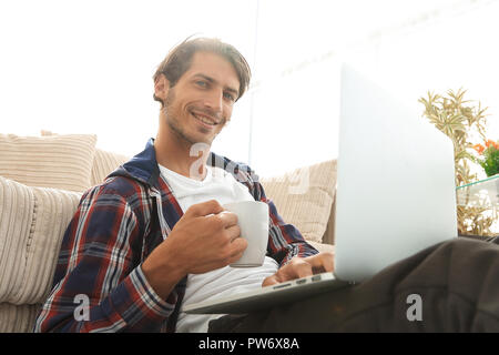 Junger Mann mit Laptop Holding eine Schale sitzen auf dem Boden in der Nähe von dem Sofa Stockfoto