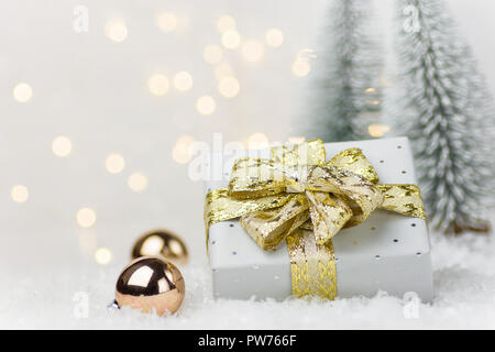 Kleine, elegante silberne Geschenkbox gebunden mit goldenen Schleife Flitter im Winter Szene im Wald mit Tannen Schnee. Weihnachten Neue Jahre präsentiert Holiday Stockfoto