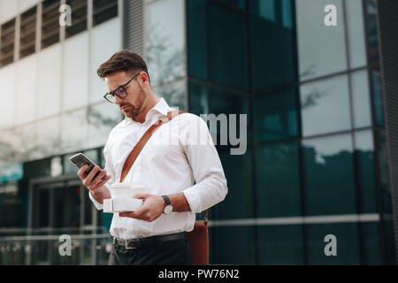 Geschäftsmann mit seinem Handy außerhalb einer Glasfassade Gebäude stehen. Mann mit zwei Tassen in der Hand über Handy ständigen Außend Stockfoto