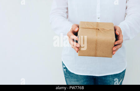 Junge kaukasier Frau in Jeans Shirt in den Händen hält, Geschenkbox in braun Handwerk Papier mit Garn gebunden. Weiße Wand. Weihnachten Neue Jahre Corporate p Stockfoto