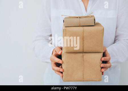 Junge kaukasier Frau in Jeans shirt hält in den Händen gestapelt Geschenkboxen in braun Handwerk Papier mit Garn gebunden. Weiße Wand. Weihnachten Neue Jahre c Stockfoto